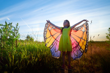 Beautiful little girl in yellow dress and in colorful bright butterfly wings in the field with green grass, yellow flowers and blue sky in a sunny summer or spring dayの写真素材