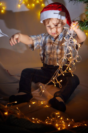 Happy little toddler boy playing with Chistmas lights in a cozy living room on New Year eve. Celebrating Xmas at home. Winter evening and male model in studioの写真素材