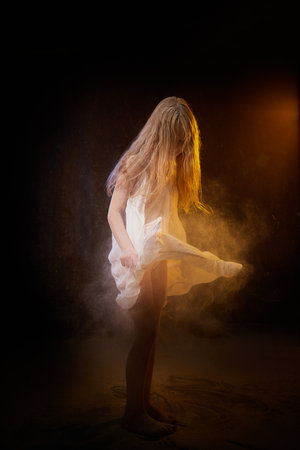 A girl in white shirt and with long hair, looking like a ghost. A young model at a photo shoot with smoke, fog, flour, dust. A little fairy girl on a dark black backgroundの写真素材