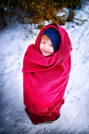 Portrait of a little frozen girl in a blanket in the snow in the forest on cold winter dayの写真素材