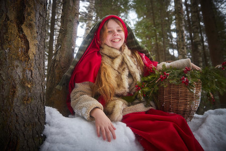 Nice cute teenager girl in old peasant clothes with hot fur coat and shawl in cold winter forest with fir trees, pines and snow on winter day. Child with basket full of green branches and red berriesの写真素材