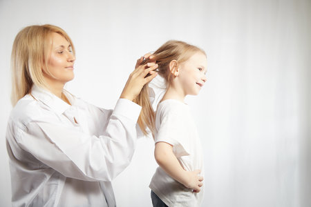 Close portrait of blonde mother and daughter where mom braids hair and makes ponytail and hairstyle for her girl on white background in studio. The concept of love, friendship, caring in the familyの写真素材