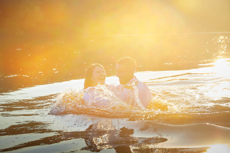 A beautiful adult couple has fun in nature in the water in a river or lake in the summer evening at sunset. A guy and a girl swim and relax outdoors in clothes in white shirts and jeansの写真素材