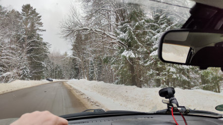 Hand of woman on the steering wheel in a car and forest and snow on the side of the road on a winter day. Woman driving car in winter travel in landscape with snow covered woodsの写真素材