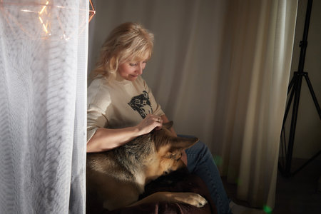 Adult mature woman of 40-60 with big shepherd dog in warm sweater. Room with calm cozy evening atmosphere with transparent curtains and soft warm light of lamps. Concept of love for animals and petsの写真素材