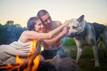 Happy wet couple relaxin, having fun and hugs with big dog near fire in camping on nature at summer sunny evening in sunset. Family or lovers have date and rest outdoor. Concept of loveの写真素材