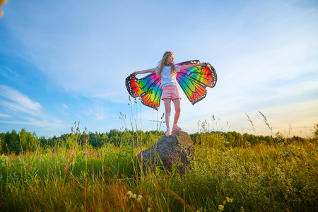 Pretty blonde girl with bright butterfly wings having fun in meadow on natural landscape with grass and flowers on sunny summer day. Portrait of a teenage child in spring season outdoors on fieldの写真素材