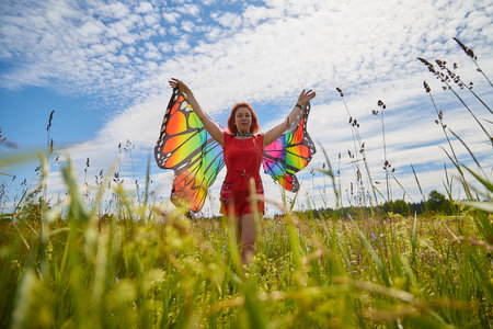 Adult girl with red hair and butterfly wings having fun and joy in meadow or field with grass, flowers on summer sunny dayの写真素材