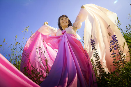 Beautiful girl in a lush pink ball gown in green field during blooming of flowers and blue sky on background. Model posing on nature landscape as princess from fary taleの写真素材