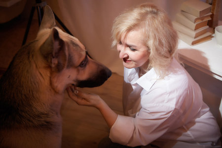 Adult mature woman of 40-60 with big shepherd dog in white shirt. Room with calm cozy evening atmosphere with transparent curtains and soft warm light of lamps. Concept of love for animals and petsの写真素材