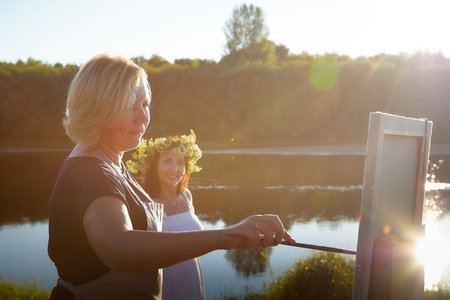Adult female artist painting picture near water of river or lake in nature and girl in white...