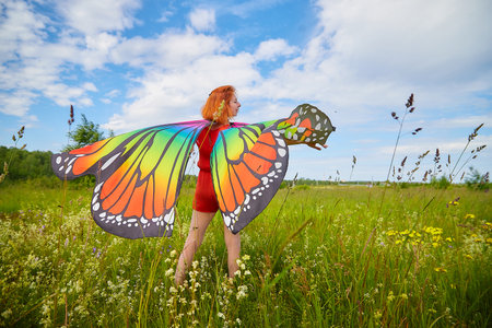 Adult girl with red hair and butterfly wings having fun and joy in meadow or field with grass, flowers on summer sunny dayの写真素材