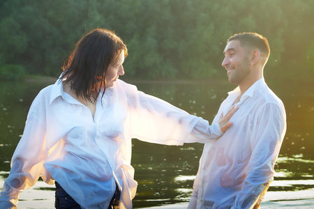 Beautiful adult couple has fun in nature in the water in a river or lake in the summer evening at sunset. A guy and a girl swim and relax outdoors in clothes in white shirts and jeansの写真素材