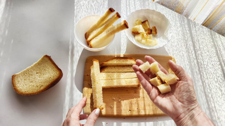 Homemade cooking and cutting of slices of diced dry white rye bread on cutting board and white plate. Hand of woman with Crackers for food, healthy eating on table in sunny day. top viewの写真素材