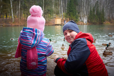 Kirov, Russia - October 23, 2022: Children at the water of the lake with floating ducks in nature on autumn day. Feeding birds and care about animalsのeditorial素材
