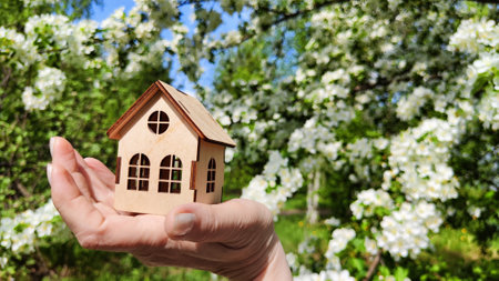 Small wooden toy house on palm of woman hand on natural background with apple flowers. symbol and concept of care, buying, selling, donating of eco friendly home. close up. soft selective focusの写真素材