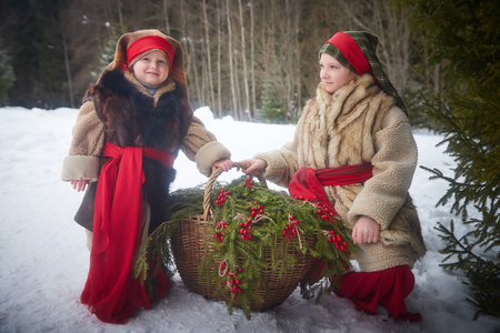 Teen and little girl in thick coat with basket of fir branches and berries in a cold winter day in forest. Medieval peasant sisters with firewood. Photoshoot in style of Christmas fairy taleの写真素材