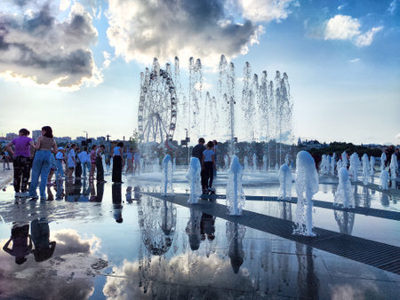 Cheboksary, Russia - June 01, 2023: Water jets of a fountain from the sidewalk and people and children cheerfully rejoice in city. Resting place for townspeople on a hot summer evening. Partial focusのeditorial素材