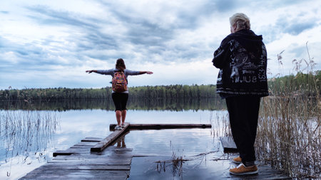 Kirov, Russia - April 29, 2023: Girls or women walk along old wooden pier with lake in background during evening and landscape featuring water, forest, clouds in sky. Tourist in natural landscapeのeditorial素材
