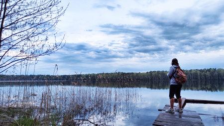 Kirov, Russia - April 29, 2023: Girls or women walk along old wooden pier with lake in background during evening and landscape featuring water, forest, clouds in sky. Tourist in natural landscapeのeditorial素材