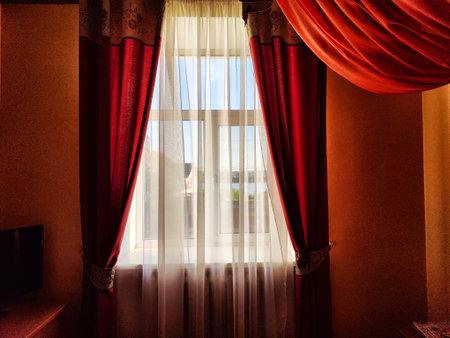 Beautiful red curtains on the window and transparent tulle fabric between them. View of daytime street. Background, texture, location, cozy room, and interior.の写真素材
