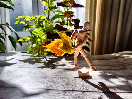 Small wooden figurine with a bouquet of autumn yellow birch leaves, bathed in sunlight, surrounded by shadows and backdrop of vegetation. Concept of autumn beauty and nature. Blurred, partial focusの写真素材