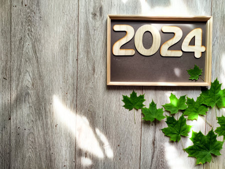 Wooden figures and numbers of the new year 2024 with frame and green maple leaves. Celebration with...