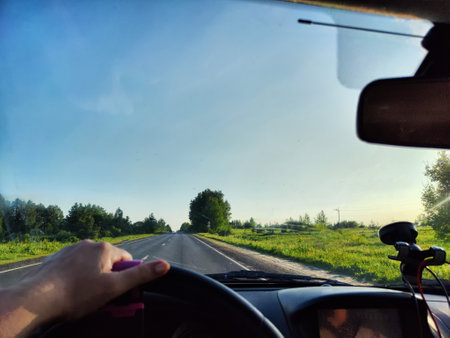 view through car windshield of natural landscape with green field, white clouds in blue sky. Hand of woman on steering wheel of car. Female traveler driving on trip or journey in spring or summerの写真素材