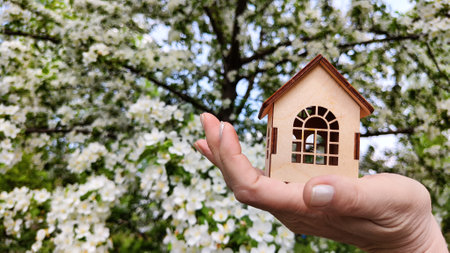 Small wooden toy house on palm of woman hand on natural background with apple flowers. symbol and concept of care, buying, selling, donating of eco friendly home. close up. soft selective focusの写真素材