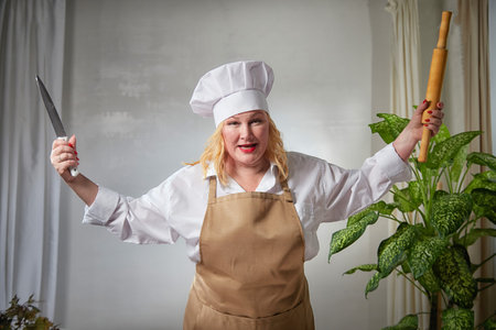 A fat evil angry funny female cook in a hat, apron and rolling pin posing and taking selfie in the kitchen. Good cooking and body positiveの写真素材