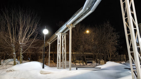Network of metal pipes, possibly for gas or water, set against a dark urban streetscape blanketed in snow, illuminated by street lamps under a winter night skyの写真素材