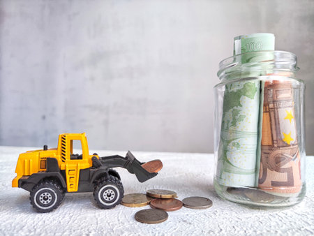 Toy Bulldozer Loading Coins Next to Jar of Cash. The concept of savings ranging from small money to...