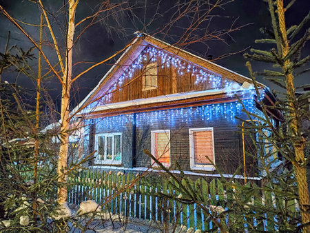 A charming wooden house adorned with blue and white Christmas lights glows amidst a snowy landscape, surrounded by treesの写真素材