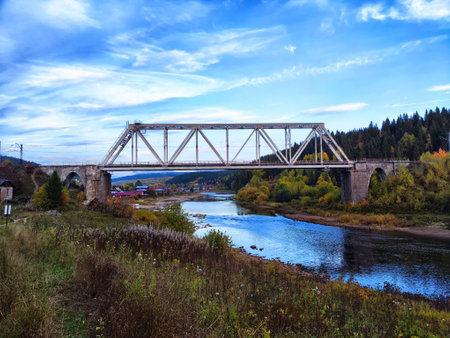 A steel bridge spans across a river in a colorful autumn forest under a clear blue skyの写真素材