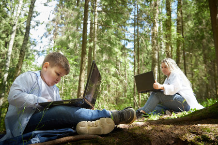 Mother and son with a laptops in forest in summer. Young boy and woman working with modern IT technologies in natureの写真素材