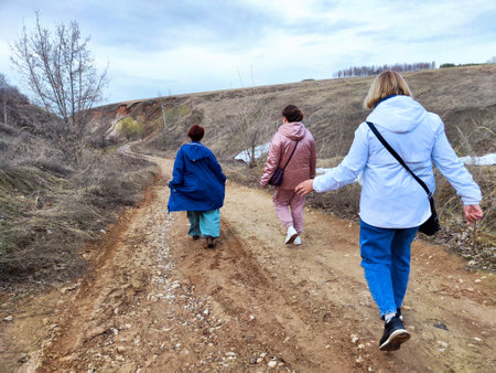 Three women hike uphill on a dirt path in a rural landscape. Field, trees, overcast sky with a few clouds. Women wear jackets, enjoying the hikeの写真素材