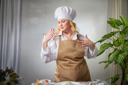 A female cook in a hat and apron poses in the kitchen and takes a selfie. Good cooking and body positiveの写真素材