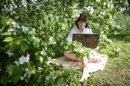 Girl Working on Laptop in Blossoming Garden. Middle aged woman using computer in white apple blossoms. Female freelancer, blogger working outdoorsの写真素材