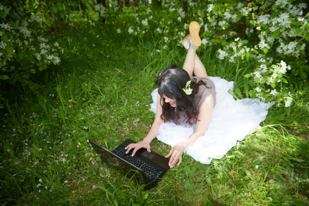Girl engages with a laptop amidst lush greenery. Freelancer Woman Working on Laptop Outdoors in a Blooming Gardenの写真素材
