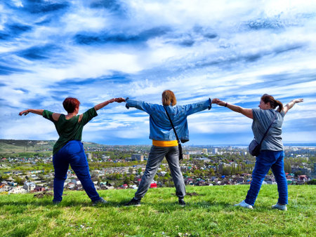 Three friends stand on grassy hilltop, arms outstretched, facing a cityscape in the distance....