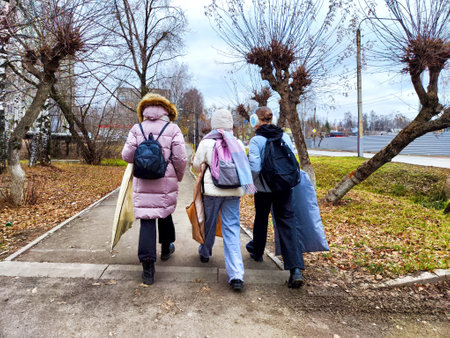 Three schoolgirls walk down a city sidewalk in autumn, carrying backpacks and other belongings. The sidewalk is lined with trees, some of which are bare, while others still have leaves. The ground is covered in fallen leaves.の写真素材