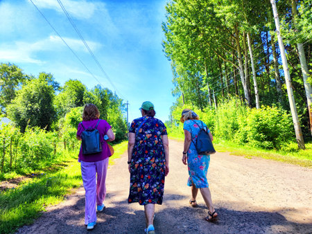 Three women in summer clothes walk along dirt road lined with green trees on a sunny dayの写真素材
