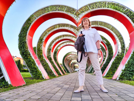 A middle-aged woman strolls through vibrant floral arches at a park during sunny dayの写真素材