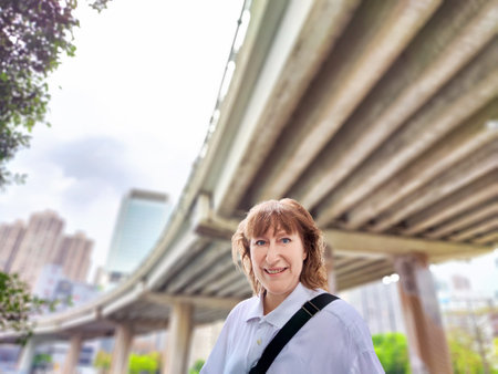 A middle-aged woman enjoys her travel experience by taking a selfie under a large bridge in Hong Kong, capturing her excitementの写真素材