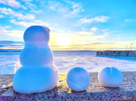 A snowman stands beside two snowballs against a sunset backdrop at a frozen lake.の写真素材