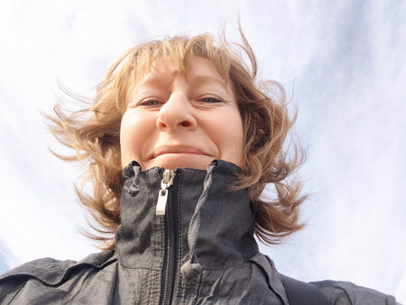 A middle-aged woman smiles broadly while traveling outdoors with a clear sky above her. The...