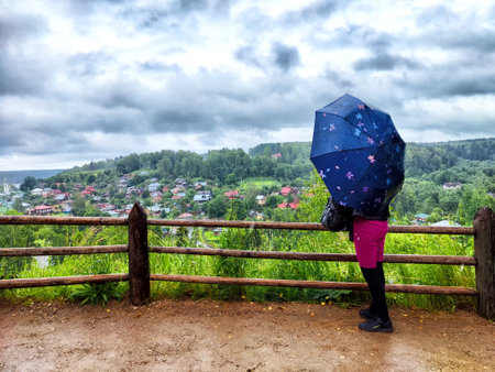 A person holding a blue umbrella stands at a lookout point, gazing at a colorful village nestled among the hills, while dark clouds hover above, suggesting rain.の写真素材
