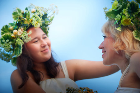 Mother and daughter in white sundresses, wreaths of flowers and bouquet on nature in evening at sunset. Family celebrate Slavic Holiday of Ivan Kupalaの写真素材