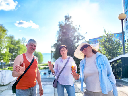 Three friends greet the camera with cheerful waves while enjoying a sunny day in a lively urban park with greenery around themの写真素材