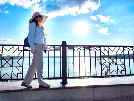 A woman strolls near the waters edge, sporting a hat and light clothing under a clear blue sky filled with sunlightの写真素材